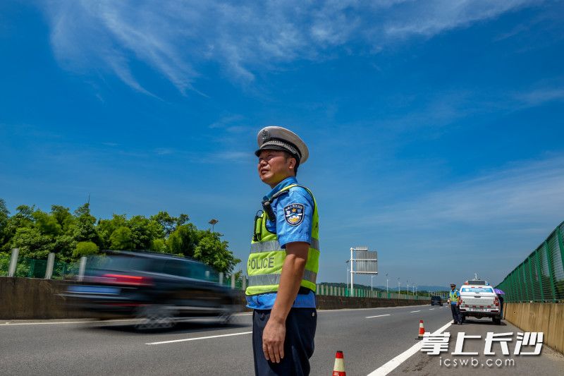 高速公路边，刘飞和袁翔顶着烈日守在故障车辆前，指挥交通，警示过往车辆，他们身旁一辆辆车快速驶过。