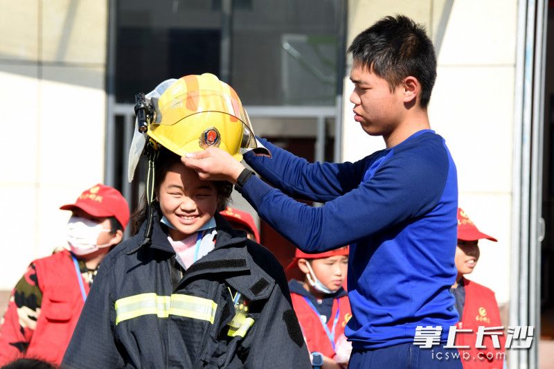 活动现场，小记者体验穿灭火防护服，消防员为其戴上消防头盔。